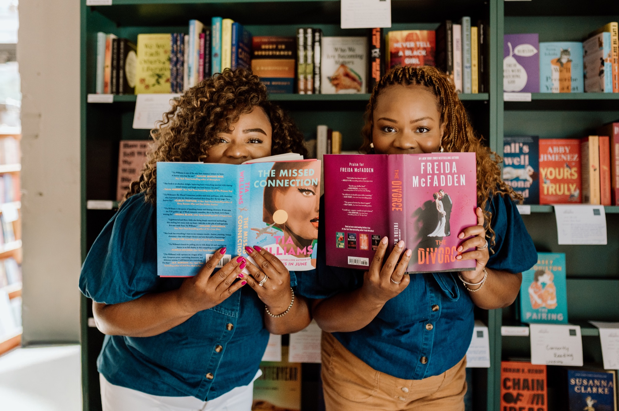 Tee and Tam peeking over thriller novels held up to their faces in a bookstore